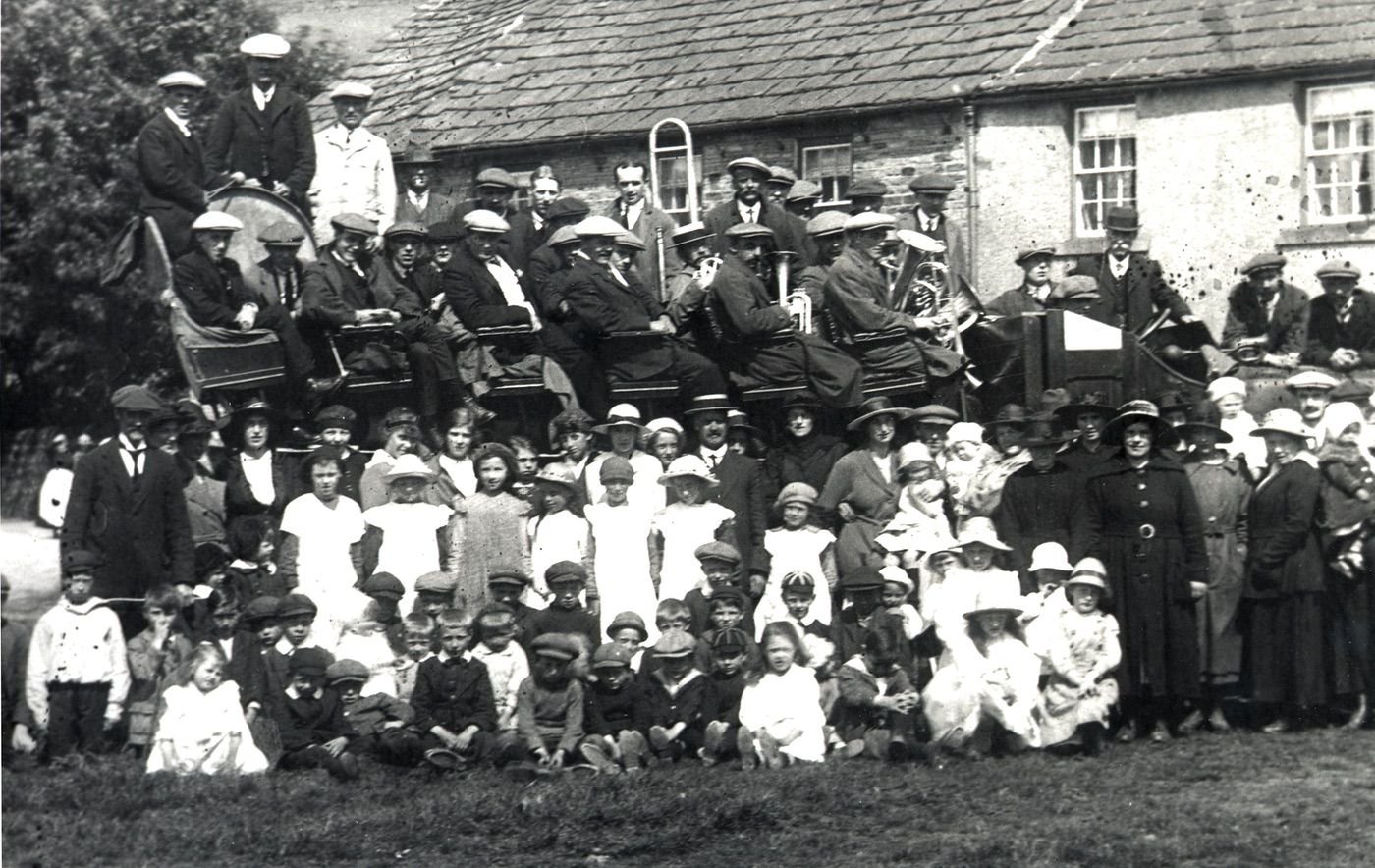 Coll. Margaret Bramwell, Meadow View, Garrigill T 381255
Collected july 2004.
Everyone's here, but confused by the visiting band and thier charabanc. The year's around 1919 and the feeling's summery so why not have ourselves photographed on Garrigill Green?
The high collared bloke standing on the left is Will Vipond- 3 folk away in the white top looks like pat Lunan's mam- the big hatted woman below the tuba is a Raine- Eric and Stewart bell are near the right front with their Wolf Cuc type caps, (popular on Alston Moor in the late 1890s)- looking straight at us in black, showing her belt buckle beneath the steering wheel is Miss Shield who had the shop- nearby at the right of the picture in a pale  wide brimmed hat is a possible Hilda Kirsopp.