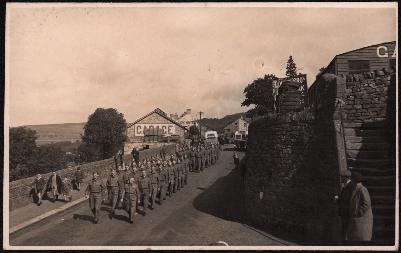 Coll. AMHS. Photographic postcard, unposted & unascribed.
Shows the LDV/Home Guard marching towrads Brewery Bridge beside Collins’ Bros garage. Two leading fiures are Tomm haldon and Fred kearton. Henderson’s garage to the L, (prior to the creation of Henderson’s tip). 5 busses inc one of Wright’s Bros of Nenthead are parked up and the Golden Lion Hotel has dropped the ‘Golden” on its gable end. The original steep steps to the Hospital Lonnen show to R.