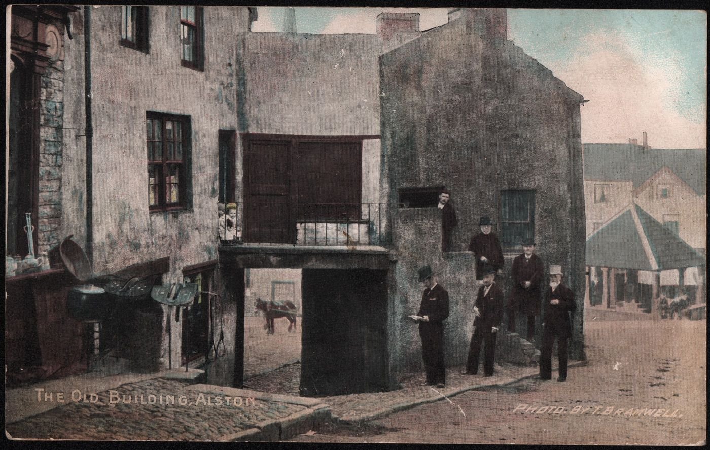 Coll. AMHS. Faded, tinted Polglase’s Series postcard version of Thomas Bramwell’s famous pre 1896 image showing some of Alston Town Council posing on the steps of a soon-to-be knocked down building.
In ca1900 the central buildings were demolished. Councillor G W  Storey, brother of Thirwall (druggist and game dealer), stands with his thumb in his waistcoat, facing his Potato Market premises. Councillor Millican, in a pale top hat, was later to become the mayor of Newcastle upon Tyne. (He was listed as a milliner but is also thought to have “handled money carefully”!). Joseph Dickinson of Lovelady Shield stands in the middle of the steps and besports short white whiskers. (Joseph was born on August 25th 1811 and died on November 26th 1895 aged 84), having been prominent in many local charities including the Fair Hill recreation ground.)
To the left is Harrison Hall’s ironmongery and saddlery- he also sold animal feeds.
Veso: , to Master Arthur Bramwell, Church View, Alston ws the message, “you will see part of Alston as it used to be.” Posted 1905.